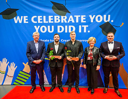 Zwei Absolventen und drei Gratulant*innen stehen vor einer Plakatwand für ein Foto