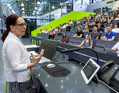 Profilansicht einer Professorin, die in einem vollen Hörsaal eine Vorlesung hält.