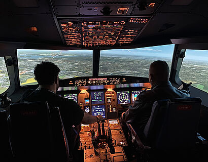 Zwei Personen sitzen in einem Flugsimulator A320 der TH Wildau und schauen aus dem Cockpit nach draußen in die virtuelle Landschaft