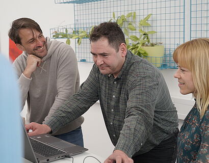 Zwei Männder und eine Frau stehen an einem Schreibtisch und schauen auf einen Laptop