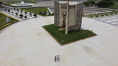 Drei Personen stehen auf einem weiten Platz vor einem monumentalen Unabhängigkeitsdenkmal, aufgenommen aus der Luft.