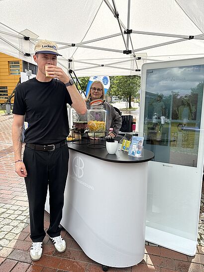 Ein Mann mit Cap trinkt aus einem Becher am Infused-Water-Stand der TK; im Hintergrund sitzt eine weitere Person hinter dem Tresen, daneben ein Banner und ein kleiner Bildschirm unter einem weißen Pavillonzelt im Freien.