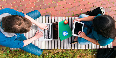 Studierende sitzen auf Bank auf dem Campus vor Laptop