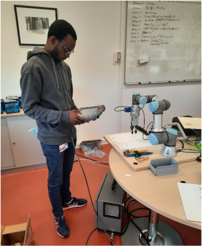 Ein junger Mann steuert mit einem Handgerät einen Roboterarm an einem Labortisch.