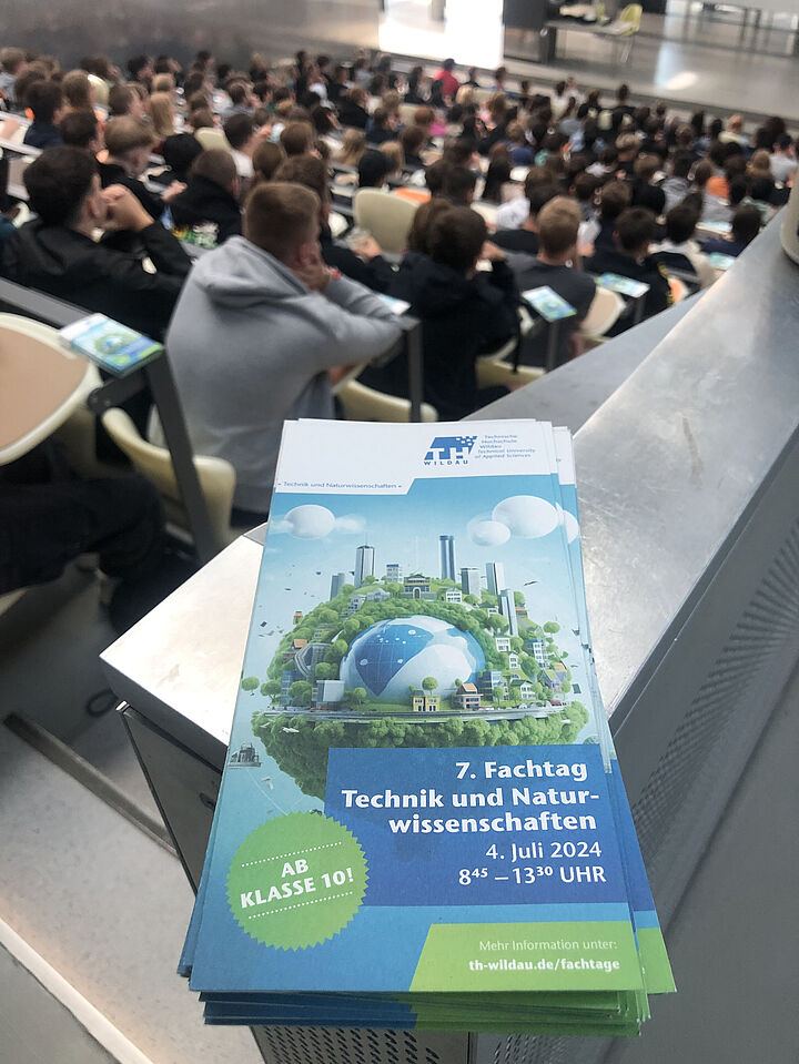 Broschüre zum Fachtag Technik der TH Wildau liegt auf einer Metallfläche und im Hintergrund sind Schülerinnen und Schüler zu sehen