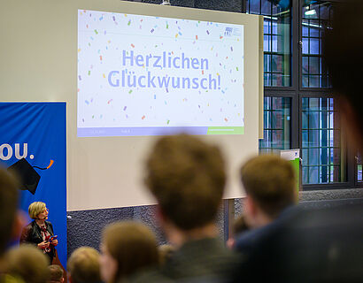 Großer Screen mit der Aufschrift "Herzlichen Glückwunsch" und Köpfe von Personen im Vordergrund
