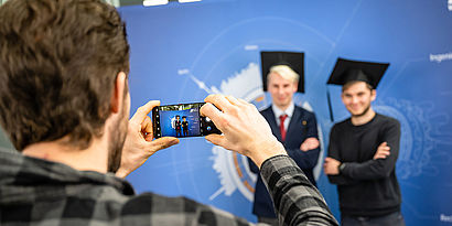 Zwei Absolventen bei der feierlichen Verabschiedung vor der blauen Fotowand mit Studierenden-Hüten werden von einem Gast mit dem Handy aufgenommen