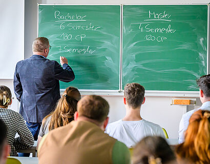 Ein Professor schreibt etwas an eine Tafel, im Vordergrund sitzen Studieninteressierte und schauen ihm zu