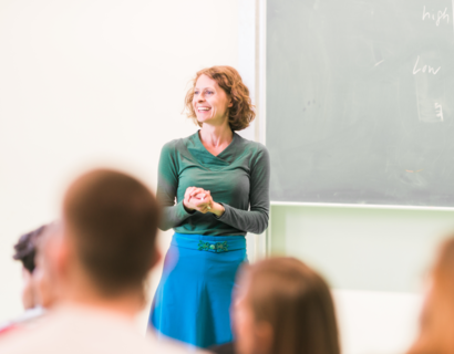 Frau Prof. Sandra Haas steht in einem Kursraum, vor einer Tafel und hält eine Vorlesung, sie lächelt die Studierenden an, trägt ein grünes Oberteil und einen türkisfarbenen Rock