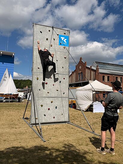 Eine Person klettert gesichert an einer grauen, rund 5 Meter hohen Kletterwand mit TK-Logo; eine zweite Person sichert von unten. Im Hintergrund stehen Zelte, ein historisches Backsteingebäude und Segelboote.