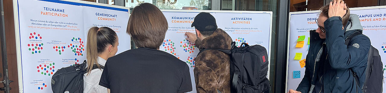 Studierende stehen vor Pinnwänden mit Plakaten und geben über bunte Klebepunkte Feedback zu verschiedenen Bereichen