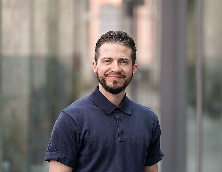 Person mit dunklem Shirt, schwarzen Haaren und Bart steht für ein Portraitbild in einer alten Industriehalle mit Betonpfeilern im Hintergrund.